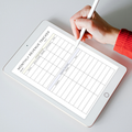 A hand holding a white stylus writes in a monthly revenue tracker spreadsheet displayed on a tablet screen, with a red ribbed sleeve visible.