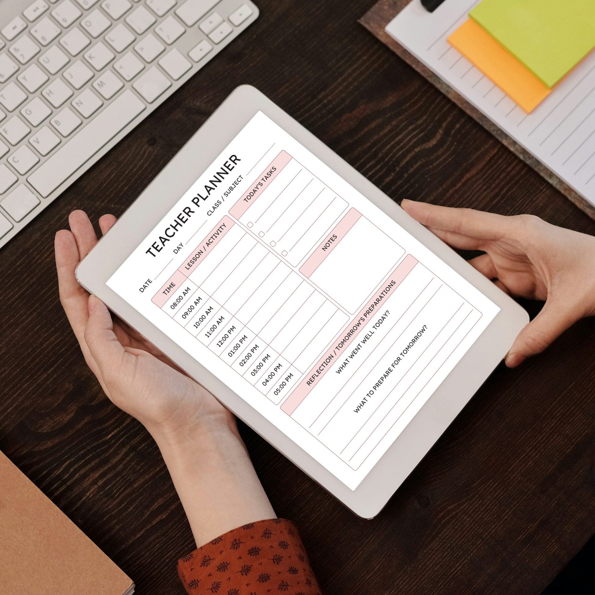 A person holds a tablet showing a digital teacher planner template on a dark wooden desk, with a keyboard, sticky notes, and a notebook nearby.